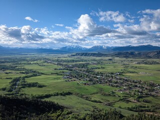 Fototapeta premium Panoramic vista of a valley town nestled amidst verdant fields and majestic snow-capped mountains under a partly cloudy sky