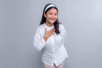 Portrait of a young Asian woman smiling gently while placing her hand on her chest, dressed in a white blouse with a headband, showing a sincere and grateful expression.