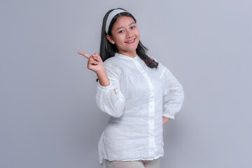 Portrait of a smiling young Asian woman in a white blouse pointing to the side with her index finger. Confident pose on grey background, perfect for marketing or promotional content.