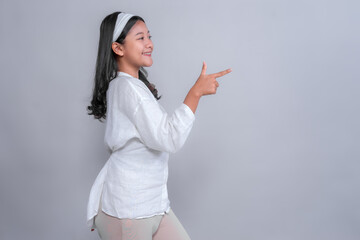 Smiling young Asian woman in white outfit making playful finger gun gesture while standing sideways. Studio shot with gray background, perfect for advertising use.