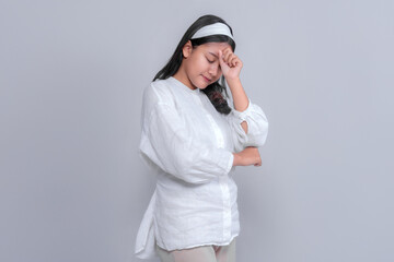 Sad young Asian woman in white outfit closing her eyes and touching her forehead with a tired expression, portraying stress or disappointment. Studio portrait, gray background.