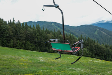 Empty chairlift glides above a green alpine meadow with pine-covered Carpathian mountains in...