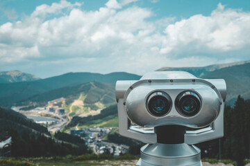 Metal binocular viewer on mountain viewpoint with scenic valley and forest in background. Clear summer day with blue sky and clouds, perfect for sightseeing and nature exploration
