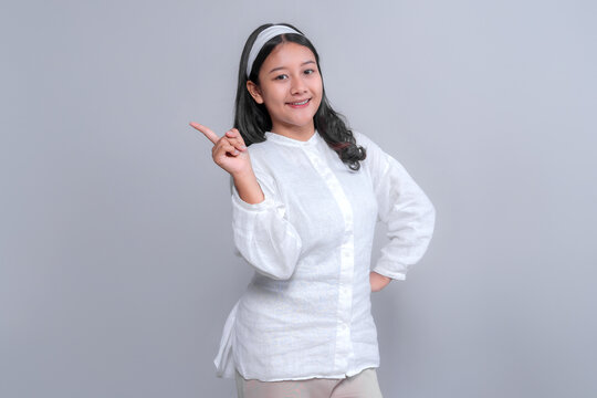 Portrait of a cheerful young Asian woman wearing a white shirt and headband, smiling confidently while pointing sideways, standing against a plain light grey background. - Powered by Adobe