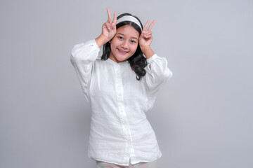 Playful young Asian woman in a white shirt and headband smiling brightly while making double peace signs near her face. Shot in studio with a clean grey background.