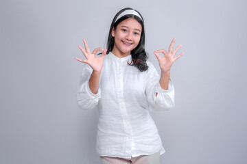 A smiling Asian woman in a white blouse and headband, making the "OK" sign with both hands. The simple gray background emphasizes her positive and friendly expression, perfect for promotional content.