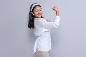 Confident Asian woman in a white blouse striking a strong pose with her fist raised, showcasing empowerment and strength. She smiles while looking back, with a neutral gray background.