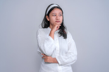 Asian woman in a white blouse thoughtfully posing with one hand on her chin, looking to the side with a curious expression. The minimalistic studio setting highlights her contemplative mood.