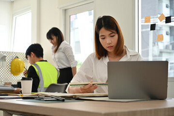 Young female engineer measuring blueprints while colleagues collaborate in the background, modern engineering office