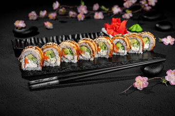 Elegant sushi roll with smoked eel and unagi sauce presented lengthwise on a textured black background with a delicate blooming sakura branch in the background. Japanese food presentation.