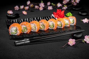 Elegant sushi roll with salmon presented lengthwise on a textured black background with a delicate blooming sakura branch in the background. Minimalist and artistic Japanese food presentation.