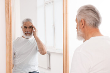 Handsome senior man looking at himself in mirror indoors