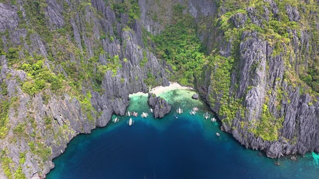 Drone view over Secret Lagoon and Jiji's Beach on Miniloc Island, Philippines, with clear waters, karst cliffs, forested terrain, white sand, and traditional boats floating in a peaceful cove
