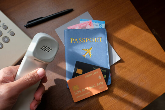 A person holding a hotel phone handset next to travel essentials including a credit card, passport, and currency on a wooden desk. Suitable for tourism, hospitality, or financial transactions