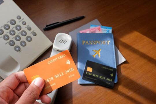 A hand holding a contactless credit card near a passport with travel money, another card, and a hotel telephone on a wooden desk.Ideal for travel, financial, international trips