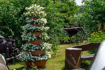 Decorative vertical arrangement of terracotta pots filled with blooming white petunias, placed on a lawn in a lush garden. Wooden bench and greenery in the background.