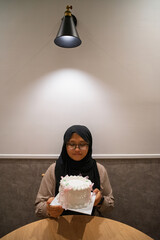 Asian muslim teenage girl wearing glasses with a vintage birthday cake sitting on the table.