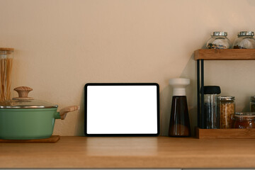 Digital tablet resting on a wooden counter surrounded by cooking utensils, jars, and pot