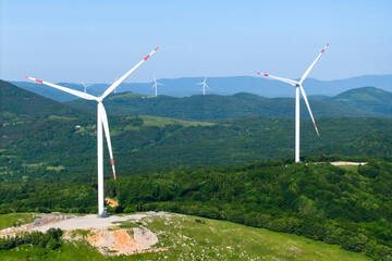 Series of white wind turbines standing on lush green hills with a backdrop of distant blue...