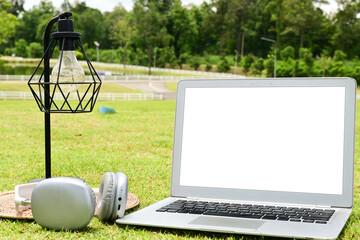 A laptop is open on a grassy field with a lamp and headphones nearby. The scene suggests a relaxed and leisurely atmosphere, possibly for someone to work or study outdoors