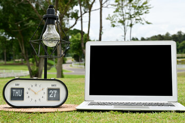 A white laptop with a black screen is placed on the green grass, with a lamp and a clock beside it.