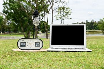 A white laptop with a black screen is placed on the green grass, with a lamp and a clock beside it.