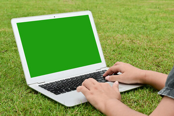 A person is typing on a laptop in a grassy field. The laptop screen is green, and the person is using a keyboard.