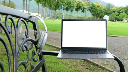 A laptop is sitting on a bench in a park. The laptop is open and the screen is blank. The bench is located in a grassy area with trees in the background.