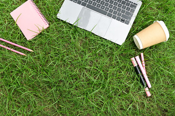 A laptop, a cup of coffee, and a few pens are on a grassy field. The scene suggests a relaxed and informal atmosphere