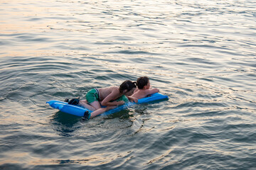 Two children swimming in sea, adrift on blue inflatable raft, accident in open water, inflatable...