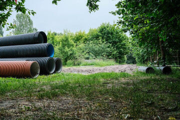 Large corrugated pipes, some with blue interiors, are stacked in a grassy area with trees and...