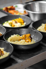 Close-up of black bowls filled with mashed potatoes and meat topped with orange sauce, served on a metal surface in a professional kitchen setting.