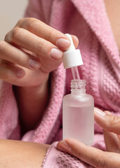 Closeup of woman in pink bathrobe opening cosmetic glass dropper bottle and holding pipette
