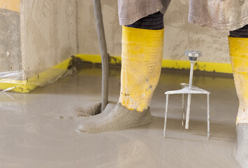 Worker wearing yellow rubber boots pouring concrete over floor in a new house construction site.