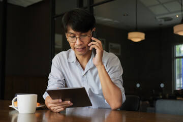 Asian man wearing glasses talking on the phone while looking at information on a tablet in his hand for work in the morning with hot coffee and croissant bakery on the wooden table at working space.