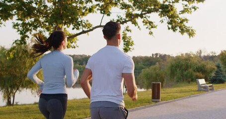 Sporty young couple jogging together along park path during sunrise. Back view of athletic woman and man in sportswear running together, enjoying healthy outdoor activity in nature. Slow motion. - Powered by Adobe