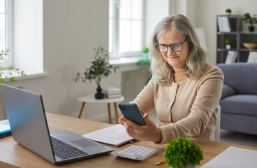 Senior woman pensioner sitting at desk working with laptop at home looking at mobile phone trough glasses having farsightedness eye disease. Mature female person using smartphone on workplace.