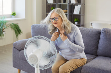 Senior woman enjoying cool air from electric fan blowing, sitting on sofa in living room interior....