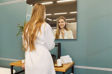 Young beautiful woman massaging face with facial massager, looking at herself in mirror, undergoing...