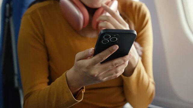 Young woman wearing casual attire and headphones around neck, sitting near airplane window while using smartphone during flight, browsing content with relaxed demeanor