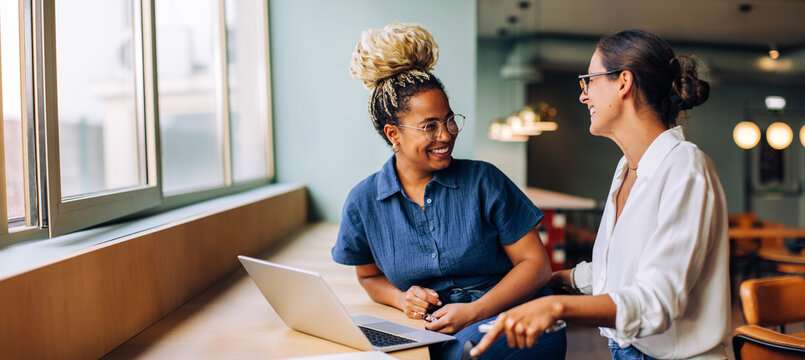 Two young women smiling and talking while working together at a laptop