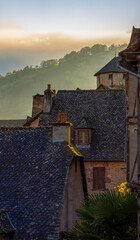 Traditional tiled roofs of stone houses in the medieval village of Conques are beautifully illuminated by the golden light of sunrise, with misty hills and a sky lit by early light.