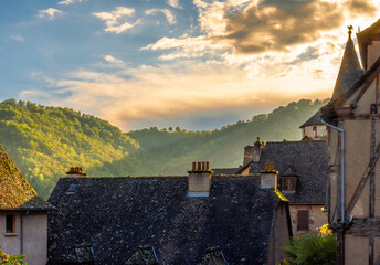 Golden hour over the rooftops of the French village of Conques, with rustic medieval houses and mountains illuminated by the sunset light under a spectacular sky with backlit clouds.