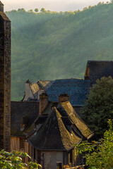 Tiled roofs of medieval houses illuminated by the light of dawn in the old French village of Conques, with mountains and morning mist in the background. France.