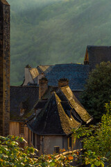 Tiled roofs of medieval village houses illuminated by the golden light of sunset in the old French town of Conques, with mountains and morning mist in the background. France.