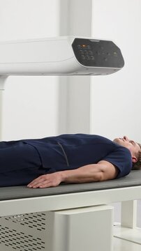A Caucasian man in his 30s lies still on a table while a modern medical scanner (DEXA) moves over his body to analyze bone density and body composition in a clinic.