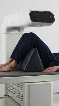 A vertical tracking shot of a male patient lying on a table with his knees supported by a positioning cushion, doing a spinal bone density DEXA scan in a modern hospital.