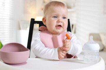 Cute little baby eating with fork on feeding chair at home