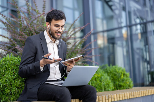 A businessman in a suit participates in a video call using a laptop and tablet outdoors, during an online meeting or conference - Powered by Adobe