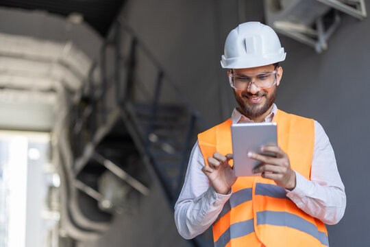 An engineer in safety gear smiles while using a tablet, inspecting a project on-site. Construction and technology integration.